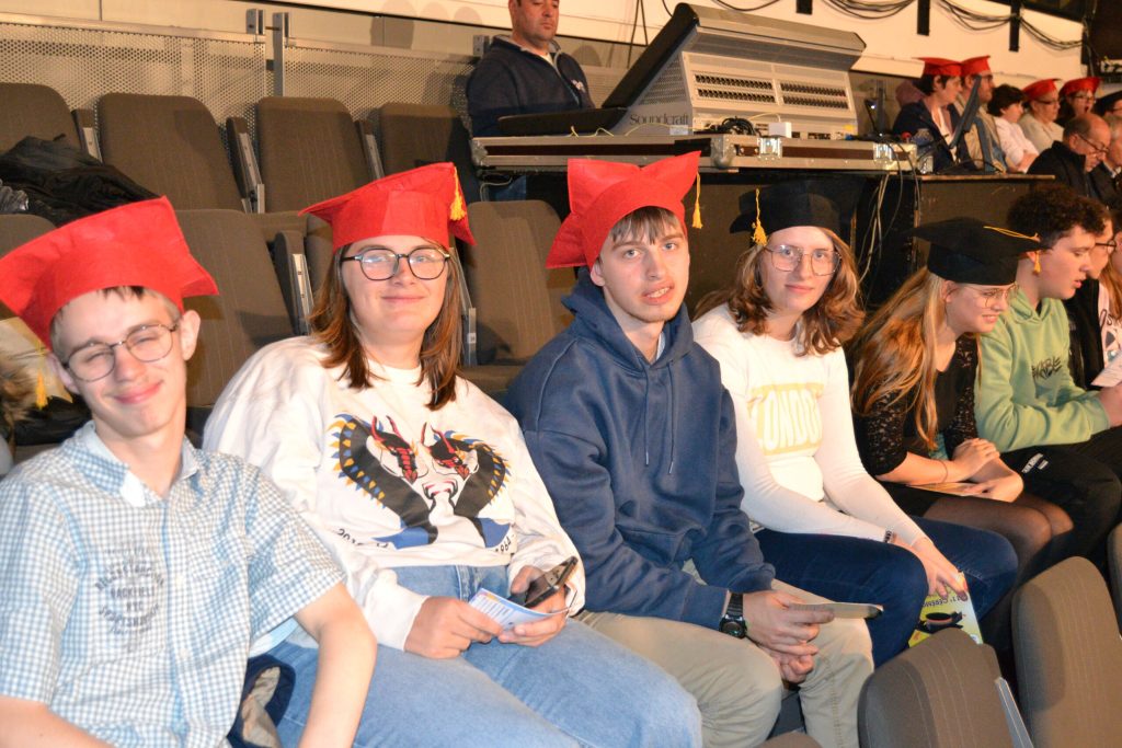 Jeunes coiffés de toques rouges et noires, souriants, assis dans les gradins lors d'une cérémonie.