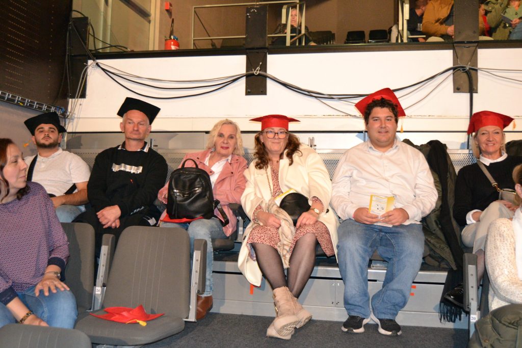 Six personnes coiffées de toques de diplômé, rouges et noires, assises dans des gradins.