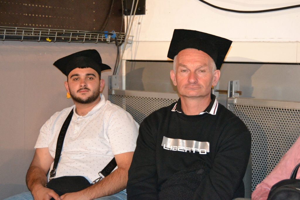 Deux hommes coiffés d'un chapeau de diplômé, assis côte à côte lors d'une cérémonie de remise de diplômes.