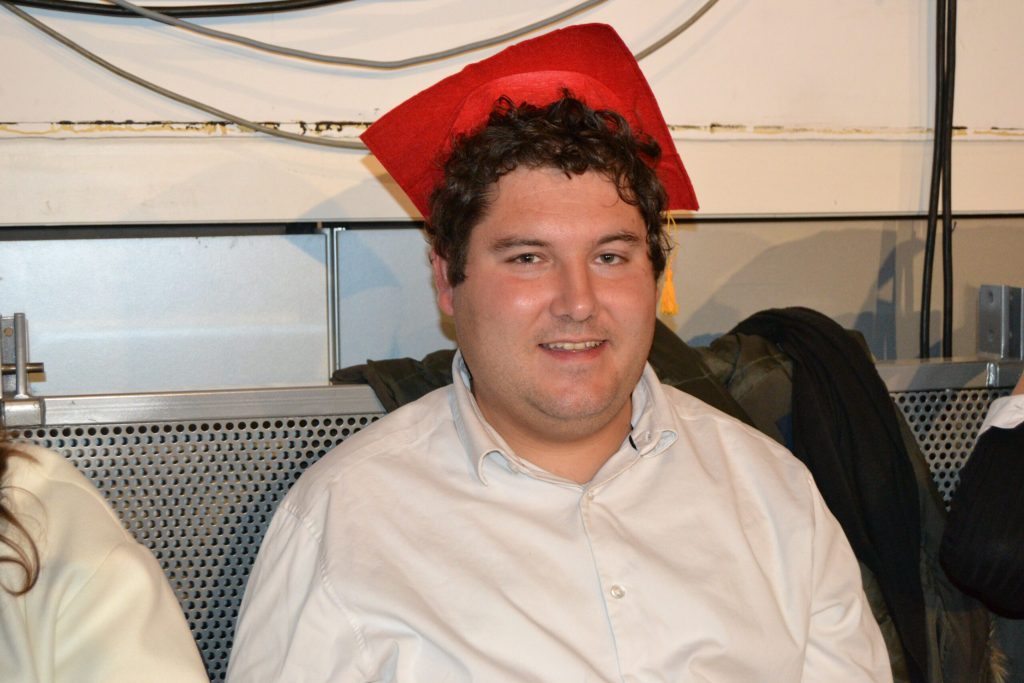 Un homme en chemise blanche coiffé d'un chapeau de diplômé rouge sourit à l'objectif.
