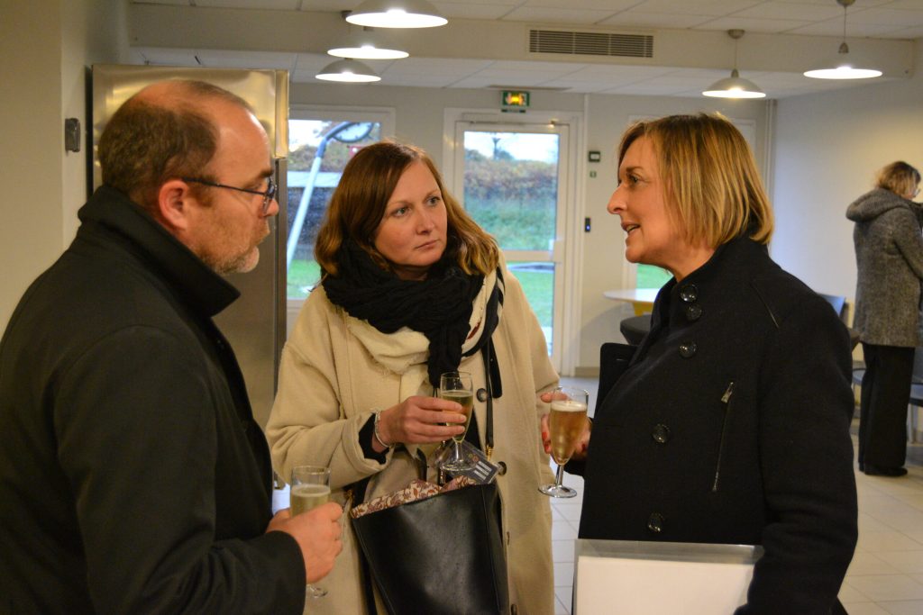 Lors d'un temps convivial, trois personnes échangent une coupe à la main dans une salle claire.