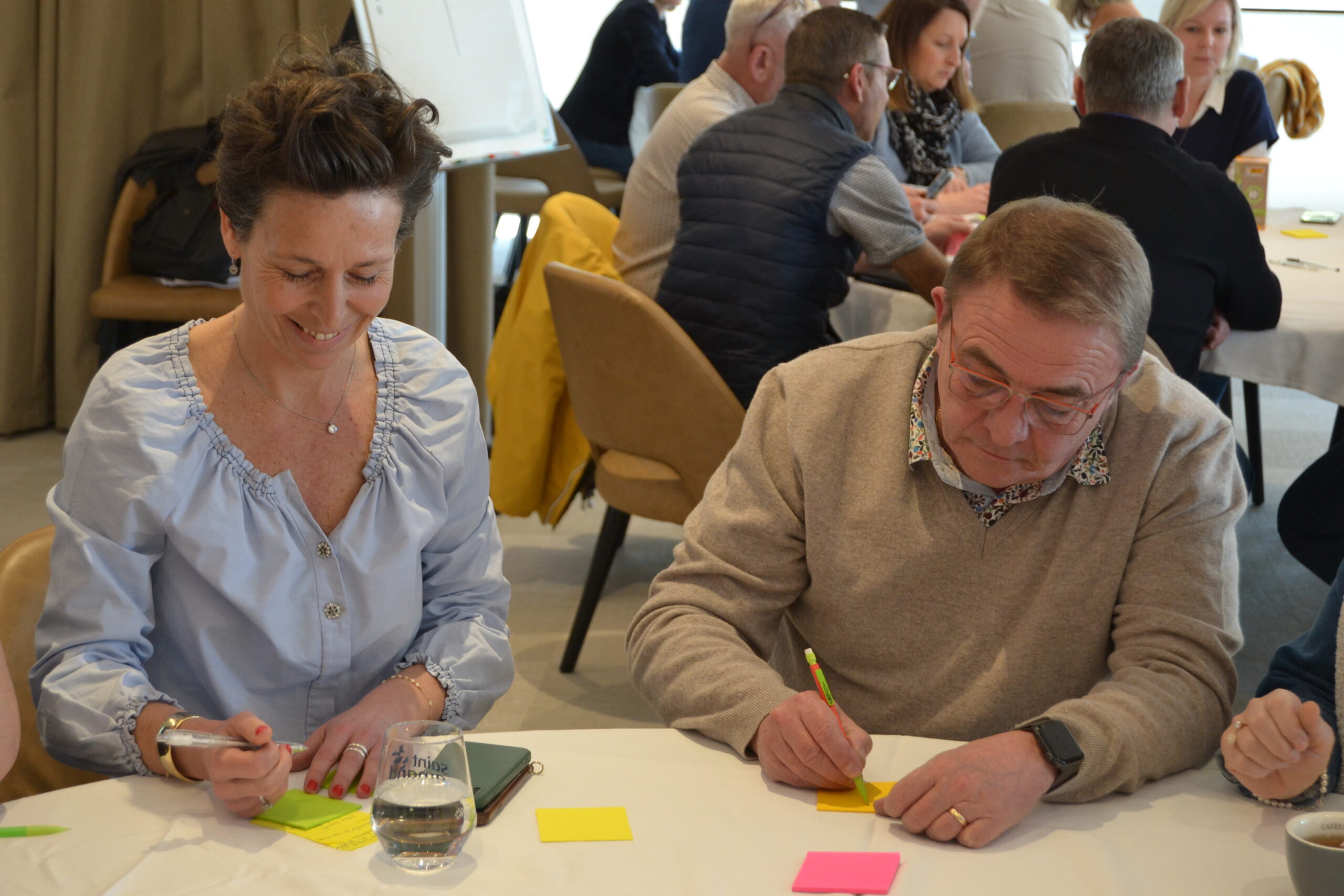 Deux participants écrivent sur des notes autocollantes lors d'un atelier, d'autres tables travaillent derrière eux.