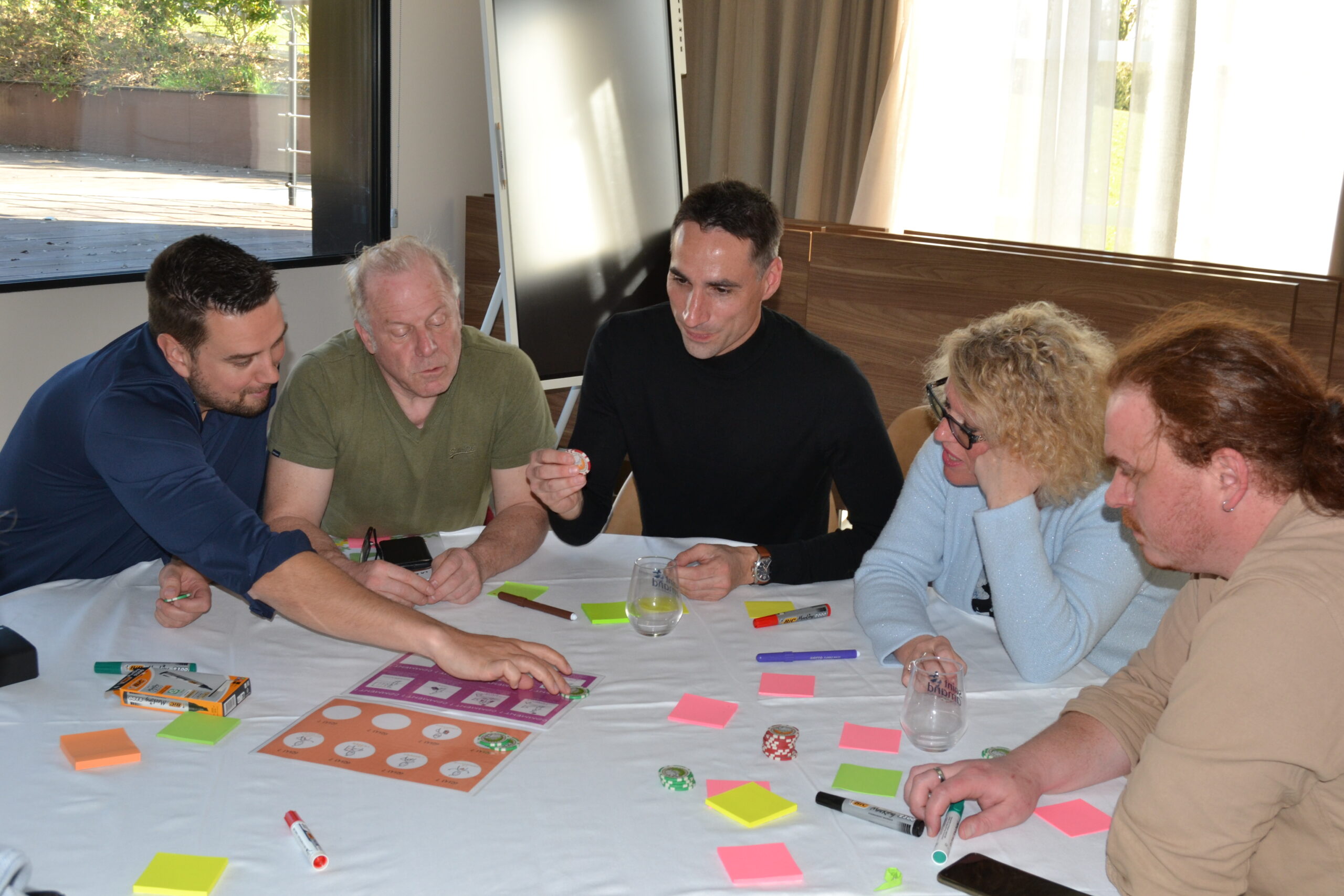 Cinq personnes réunies autour d'une table participent à un atelier avec plateau de jeu, jetons et post-it.
