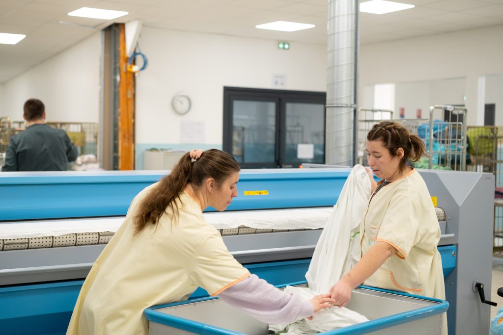 Deux salariées en blouse plient du linge à la sortie d'une calandre, dans une blanchisserie industrielle.