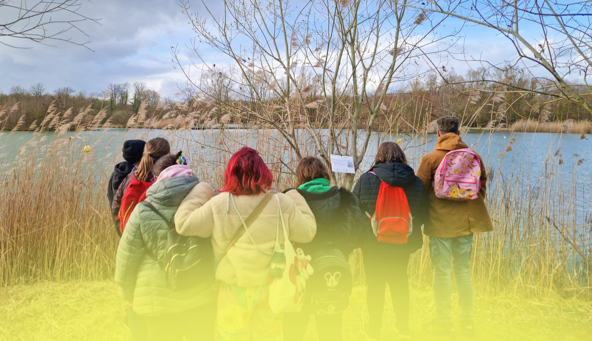 De dos, un groupe de jeunes avec sacs à dos observe un étang bordé de roseaux lors d'une sortie nature.