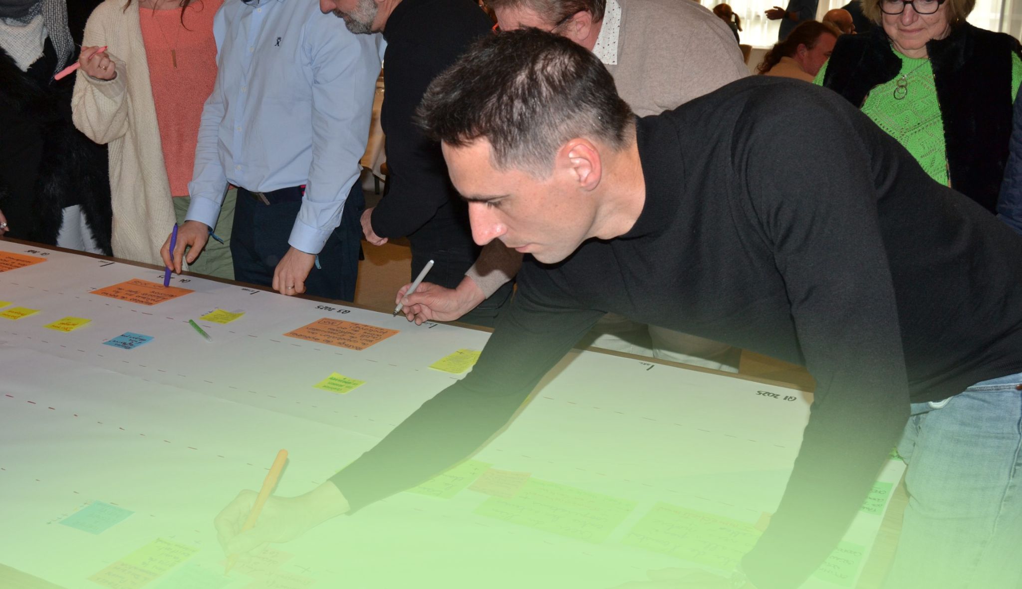 Penché sur une frise chronologique déroulée sur la table, un participant note une idée parmi les post-it, en atelier.