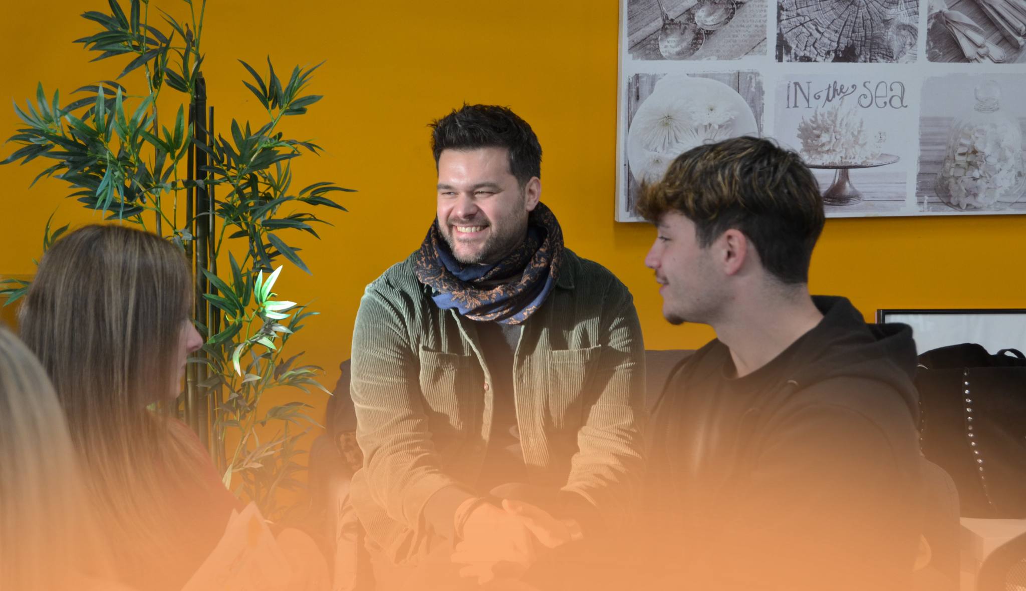 Trois jeunes adultes échangent, assis dans une pièce aux murs jaunes ; l'un d'eux sourit largement.
