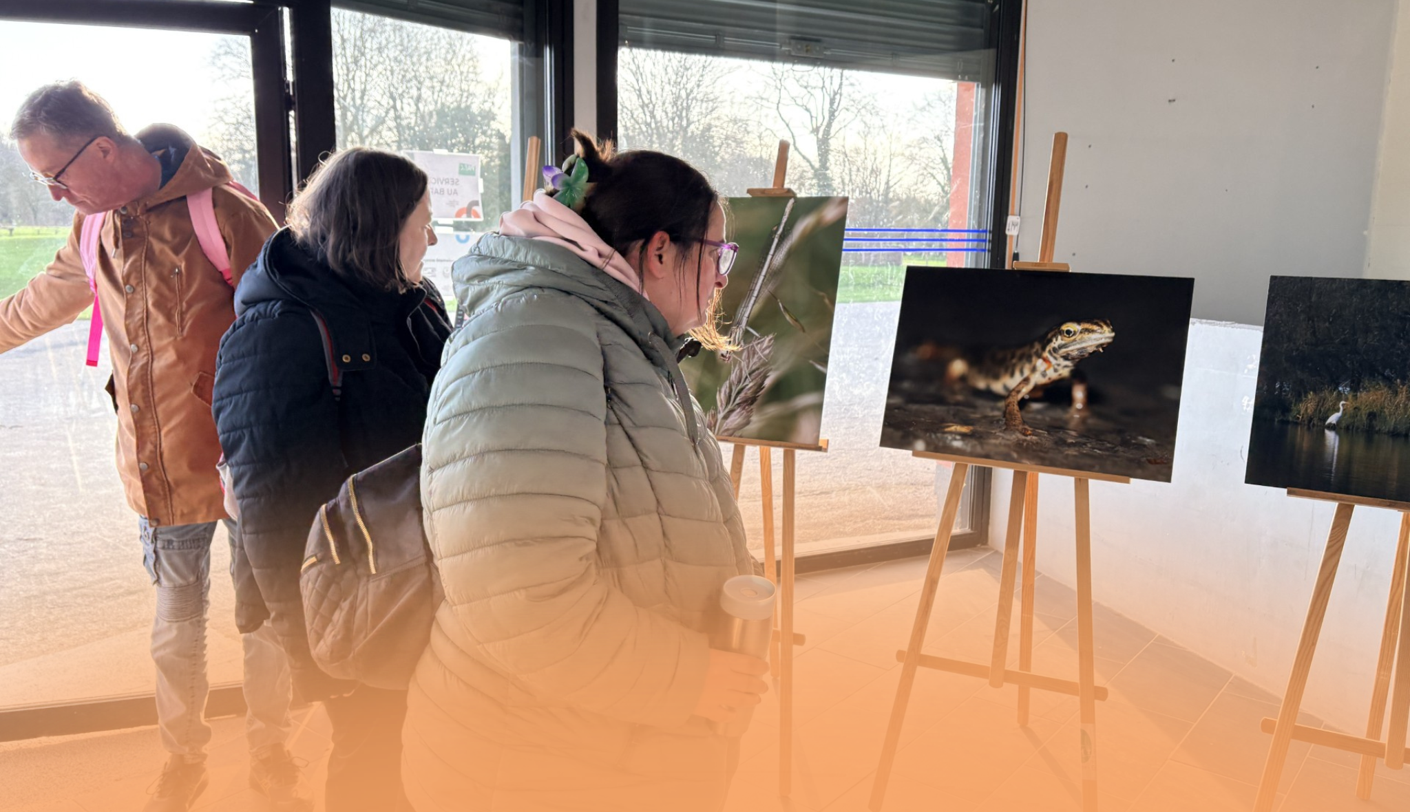 Trois visiteurs observent des photographies animalières posées sur des chevalets, près d'une baie vitrée.