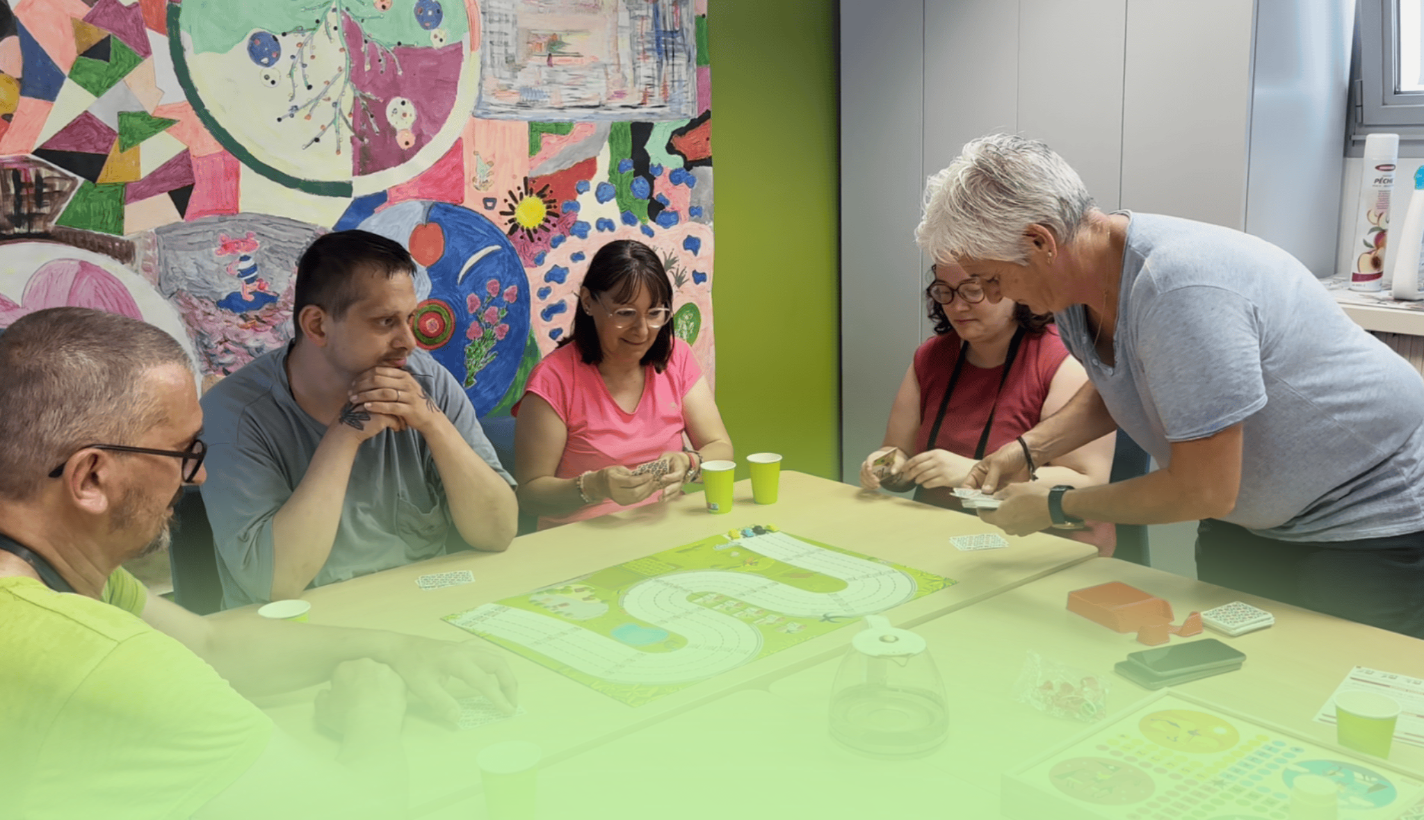 Autour d'une table, des adultes jouent à un jeu de plateau tandis qu'une animatrice debout distribue les cartes.