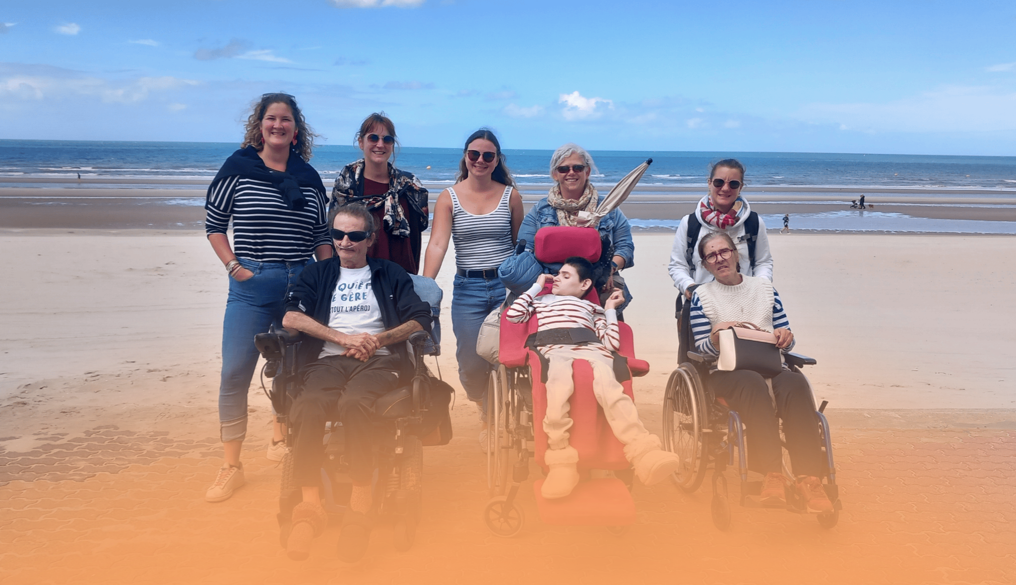 Sur une plage face à la mer, un groupe pose : cinq personnes debout et trois personnes en fauteuil roulant.