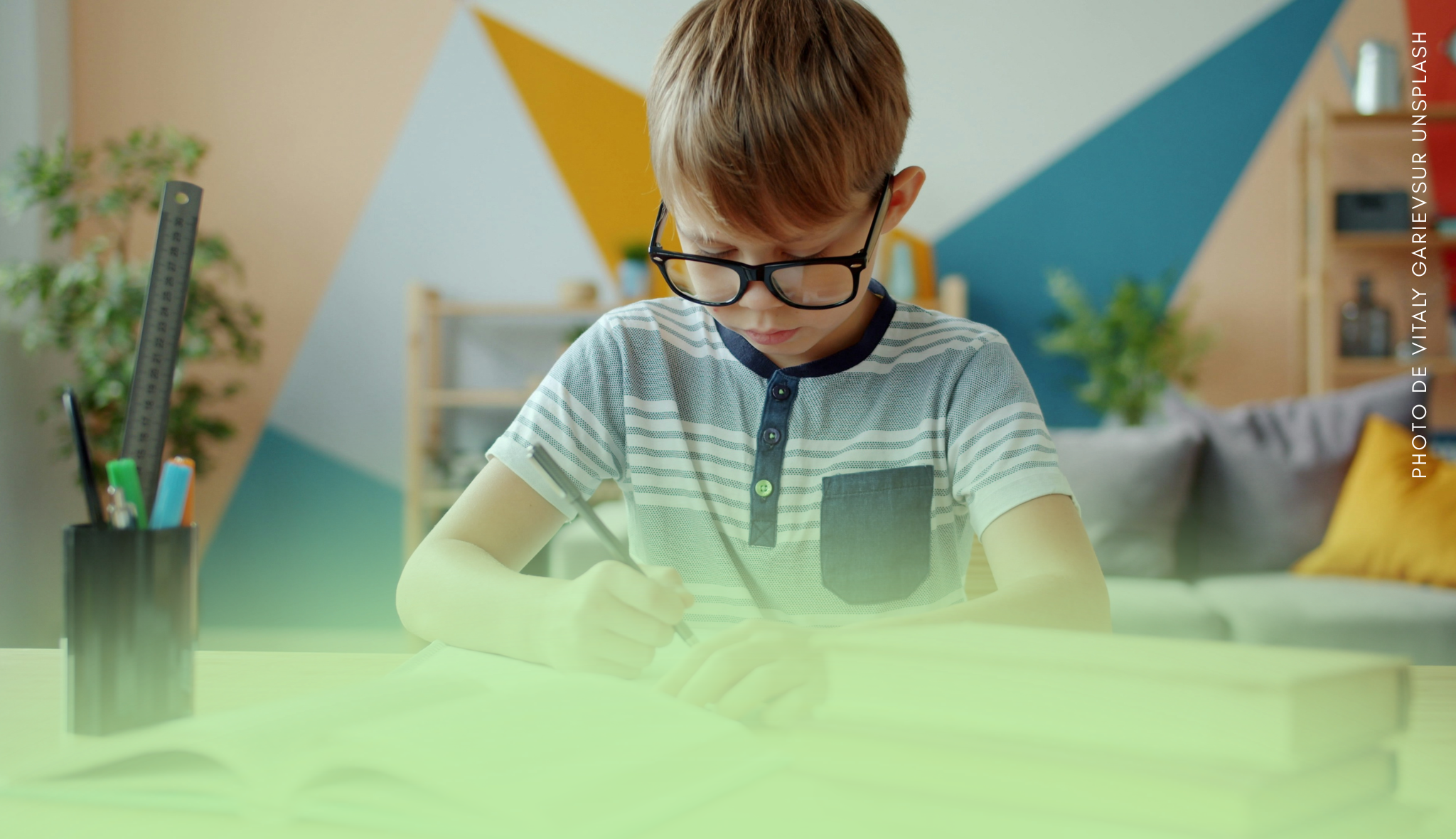 Un enfant à lunettes, concentré, écrit avec un stylo à une table dans une pièce lumineuse.