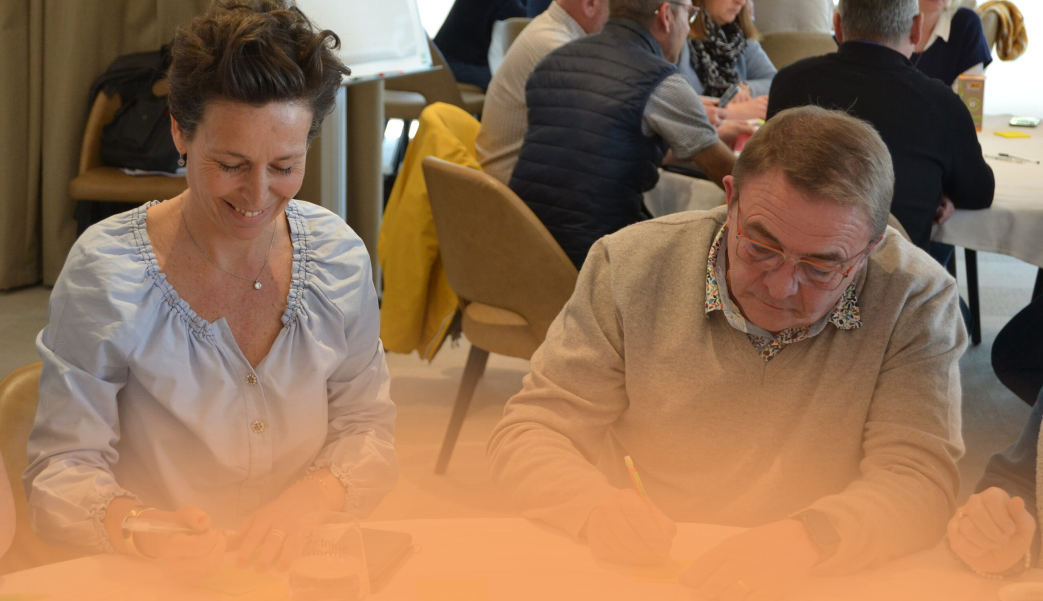 Deux participants prennent des notes côte à côte, lors d'un séminaire réunissant plusieurs tablées.