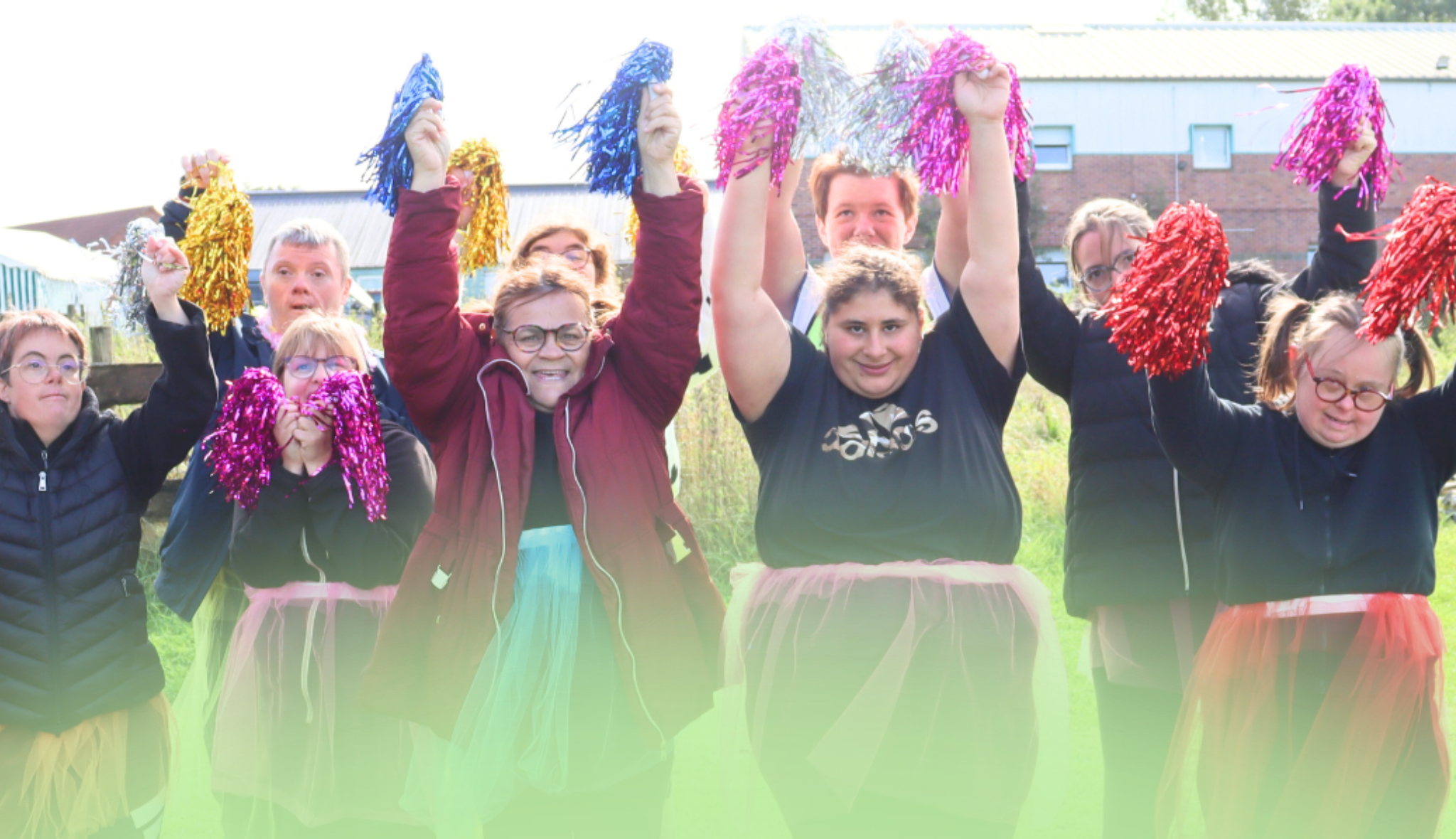 Groupe en jupes de tulle brandissant des pompons colorés, bras levés, dans un jardin devant des bâtiments.