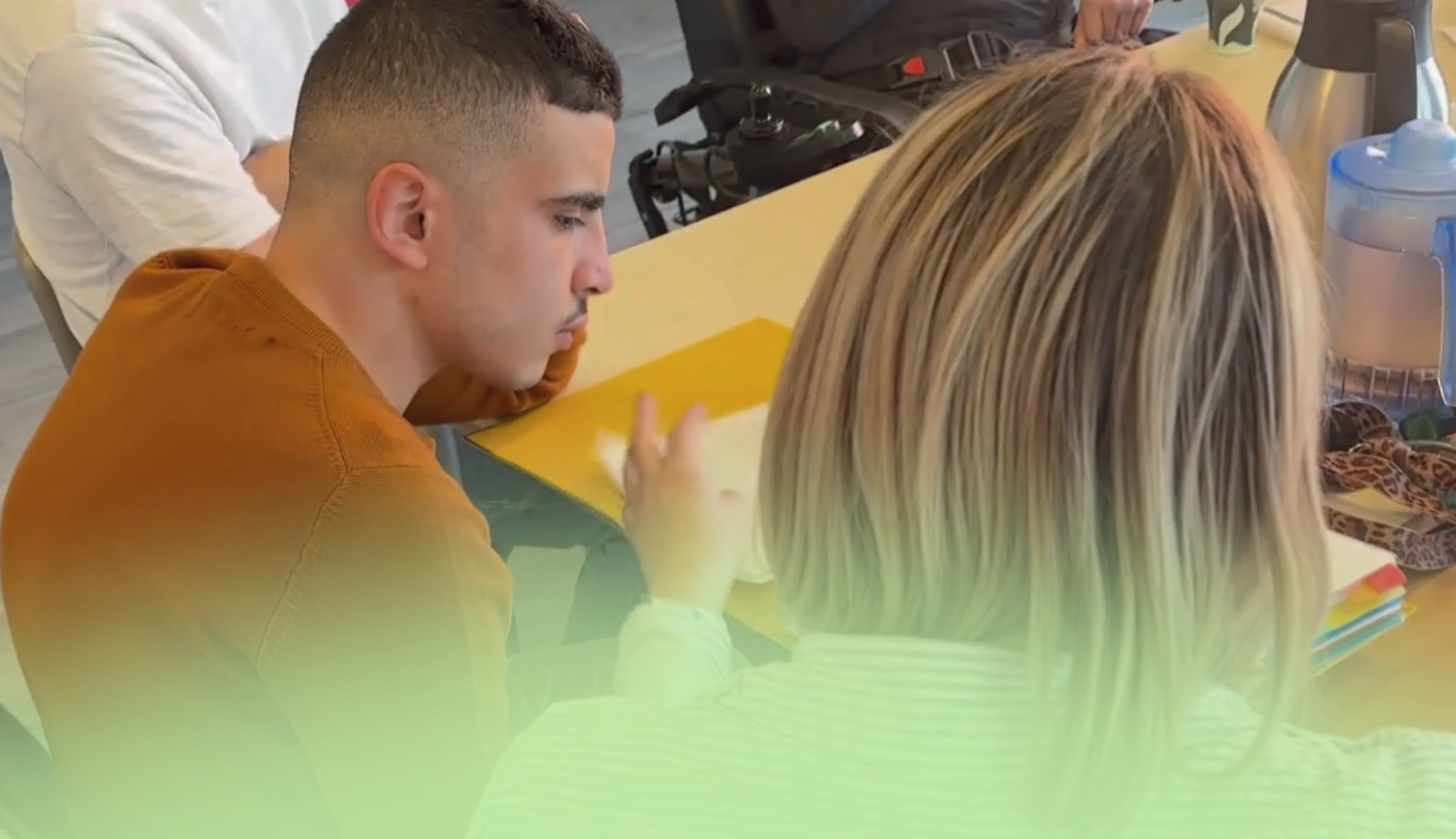Attablés face à face, un jeune homme et une professionnelle travaillent sur une feuille de couleur posée entre eux.