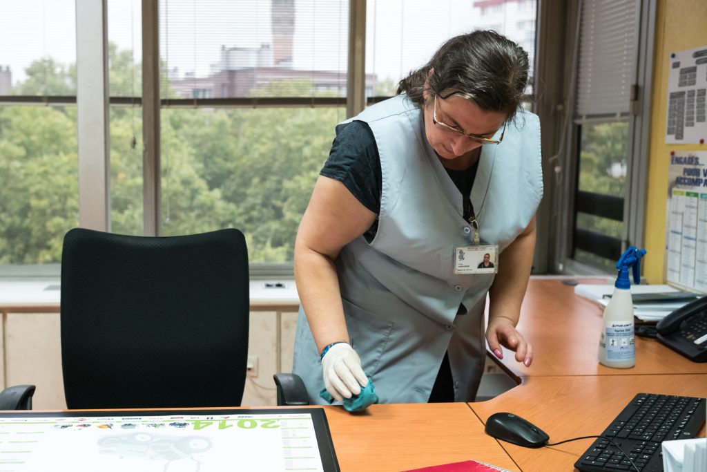 Une professionnelle en blouse et gants désinfecte le plan de travail d'un bureau équipé d'un ordinateur.